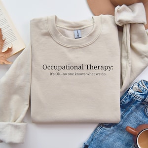 May include: A beige crewneck sweatshirt with the text "Occupational Therapy: It's OK-no one knows what we do." The sweatshirt is folded and placed on a surface with a book and jeans.