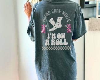 Puede incluir: Camiseta de color verde azulado oscuro con un gráfico en la espalda que dice "Wound Care Nurse" en un círculo, con una imagen de un calcetín y tijeras. Debajo del círculo, el texto dice "I'M ON A ROLL" en blanco, con un borde a cuadros.