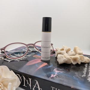 May include: A black and white roll-on bottle with a white label that says "Palamede" and "Novel Fragrance". The bottle is sitting on a book with the words "The Ninth" on the cover.