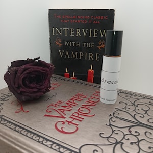 May include: A still life featuring a dried burgundy rose, a novel, and a perfume bottle. The book, "Interview with the Vampire," is displayed with lit candles. The perfume bottle is labeled "Armand Novel Fragrance."