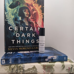 May include: A stack of four books with different titles and authors. The top book is "Certain Dark Things" by Silvia Moreno-Garcia. The second book is "Buffy the Vampire Slayer" Season One. The third book is "The Queen of the Damned" by Anne Rice. The bottom book is "The Gilda Stories" by Jewelle Gomez. A small bottle of perfume is sitting on top of the first book.