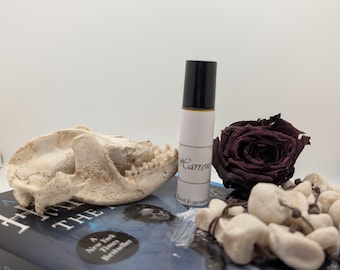 May include: A small, white, roll-on bottle with a black cap and a white label that says "Carrow" and "Novel Fragrances". The bottle is sitting on a book with a blue cover and the title "Harrow the Ninth" in white letters. There is a dried rose and a white rock next to the bottle.