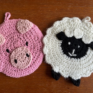 May include: Two crocheted pot holders, one shaped like a pig and the other shaped like a sheep. The pig is pink with black eyes and a black snout. The sheep is white with a black face and black legs.