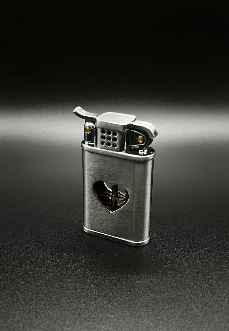 May include: A silver lighter with a heart-shaped cutout on the front. The lighter has a brass wick and a black wick cover.