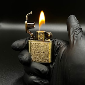 Peut inclure: Un briquet doré avec un motif complexe représentant une croix et les mots "S.P.Q.R." et "INRI". Le briquet est allumé et la flamme est visible.