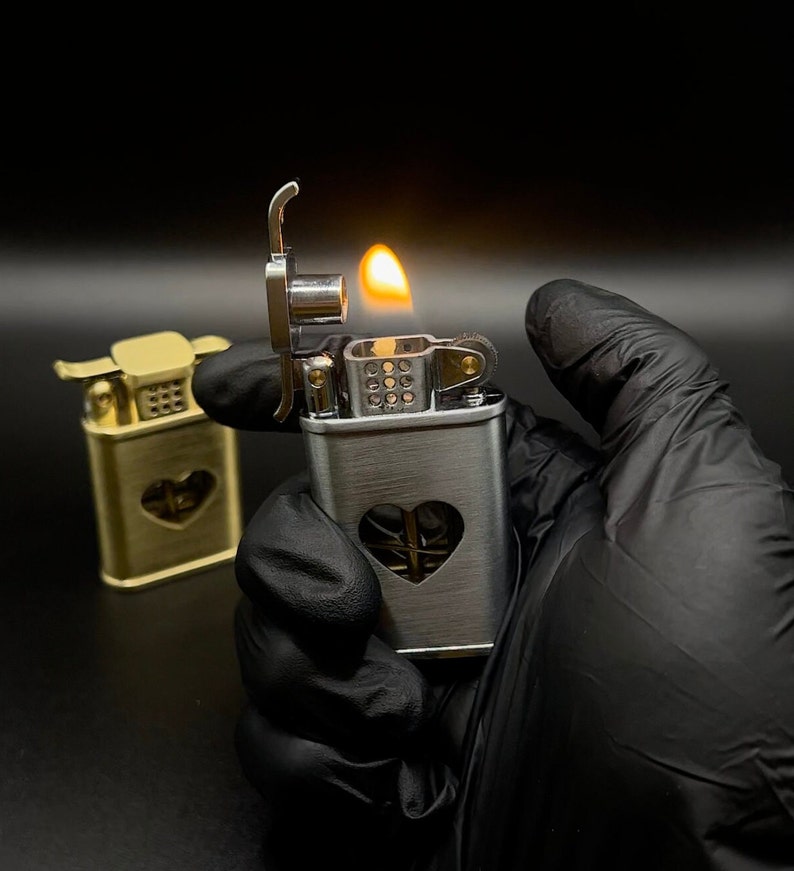 May include: A silver lighter with a heart-shaped cutout and a flame coming out of the top. The lighter is being held in a black gloved hand.