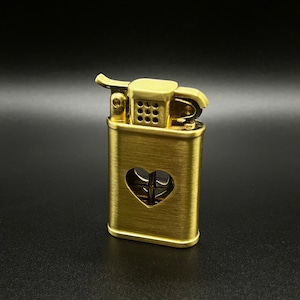 May include: A gold-colored lighter with a heart-shaped cutout on the front. The lighter has a brushed metal finish and a small, round wick.