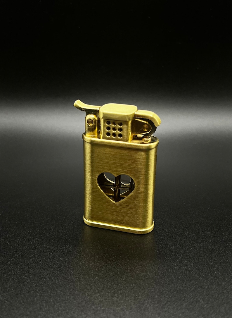May include: A gold-colored lighter with a heart-shaped cutout on the front. The lighter has a brushed metal finish and a small, round wick.