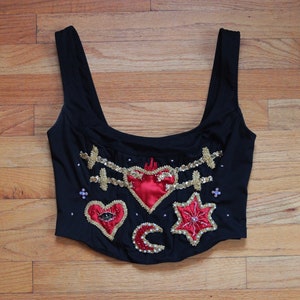 May include: Black corset top with decorative embroidery. Features include a red satin heart, a red star, and a red heart with an eye. Gold and silver embellishments and a crescent moon complete the design.