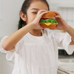 以下が含まれることがあります： 若い人が、かぎ針編みで作ったハンバーガーのおもちゃを持っています。ハンバーガーは、茶色、黄色、緑の糸で作られています。