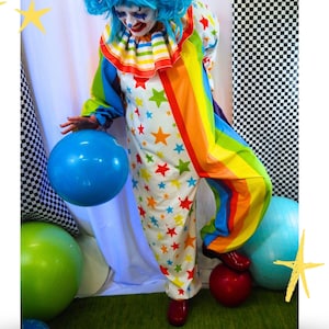 Op de afbeelding: Een clownskostuum bestaande uit een witte jumpsuit bedekt met veelkleurige sterren, een regenboogkleurige ruchekraag en lange mouwen met regenboogstrepen. De outfit bevat een felblauwe pruik met haarstrikken en rode schoenen, samen met een blauwe ballon.