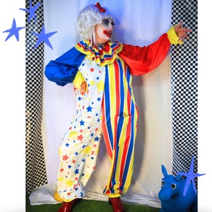 Op de afbeelding: Een clownskostuum bestaande uit een witte jumpsuit met een patroon van rode en gele sterren aan de ene kant en verticale rode, gele en blauwe strepen aan de andere kant. Het bevat een ruchekraag, lange mouwen met gele boorden en een witte pruik met een rode strik.