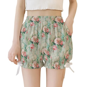 May include: Light green shorts featuring a floral pattern with pink roses and white blossoms. The shorts have an elastic waistband, a loose fit, and white ribbon ties at the bottom of each leg opening, creating a gathered, ruffled effect.