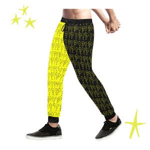 Puede incluir: Pantalones deportivos con un diseño dividido: una pierna es de color amarillo brillante con un patrón de signos de interrogación repetidos, la otra es negra con el mismo patrón de signos de interrogación amarillos. Los pantalones se usan con zapatillas negras.