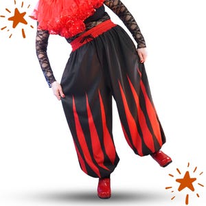 Puede incluir: Pantalones negros holgados con un patrón de llamas de color rojo vibrante que se extiende desde el dobladillo hacia arriba. Los pantalones tienen una cintura elástica con un cordón negro, creando una silueta fruncida en la cintura y los tobillos.