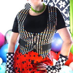 May include: A black and white striped vest with a black shirt and red star-patterned skirt. The outfit includes black and white checkered arm warmers and a green beaded necklace. The background features colorful exercise balls and a black and white floral pattern.
