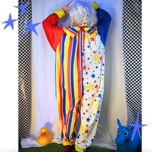Op de afbeelding: Een clown-jumpsuit met een gespleten ontwerp, waarbij de ene kant verticale rode, gele en blauwe strepen heeft en de andere kant een witte achtergrond met een patroon van kleine rode, blauwe en gele sterren, compleet met een kraag.