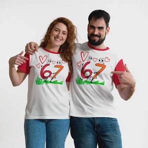 Op de afbeelding: Witte T-shirts met rode mouwen en een rode kraag. De shirts hebben een cartoonontwerp van de cijfers 6 en 7 die elkaars hand vasthouden, met harten en gras. Een man en een vrouw dragen deze shirts.