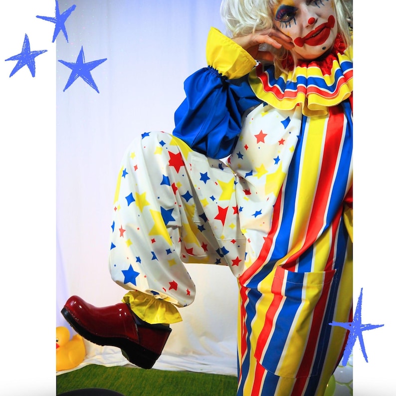 Op de afbeelding: Een clownskostuum bestaande uit een wit bovenstuk met blauwe mouwen en een gele ruchekraag, gecombineerd met een witte broek bedekt met rode en blauwe sterren. Het onderste gedeelte heeft verticale rode, gele en blauwe strepen. Een rode klomp is zichtbaar.