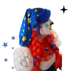 Puede incluir: Un payaso con maquillaje blanco, acentos azules y una nariz negra. El payaso lleva un sombrero azul con estrellas amarillas y trenzas rojas con mechones azules. El atuendo del payaso incluye un cuello rojo y una parte superior blanca con estampado floral.