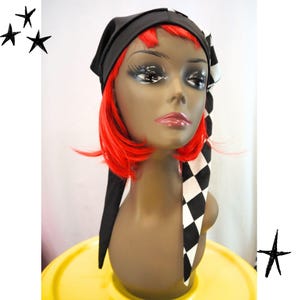 May include: A black headscarf with a long, pointed tail featuring a black and white chequered pattern. The headscarf is worn on a mannequin head with short, bright red hair, styled with long, dark eyelashes and glossy pink lips.