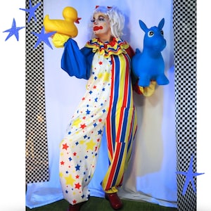 Op de afbeelding: Een clown in een witte jumpsuit met rode, gele en blauwe sterpatronen aan de ene kant en verticale rode, gele en blauwe strepen aan de andere kant. De clown heeft wit haar, rode schmink en houdt een gele badeend en een blauw ezel-speeltje vast.