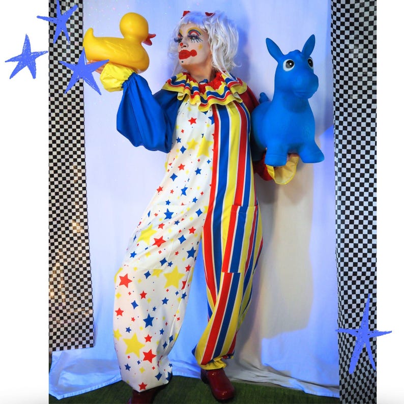 Op de afbeelding: Een clown in een witte jumpsuit met rode, gele en blauwe sterpatronen aan de ene kant en verticale rode, gele en blauwe strepen aan de andere kant. De clown heeft wit haar, rode schmink en houdt een gele badeend en een blauw ezel-speeltje vast.