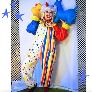 Op de afbeelding: Een clownskostuum bestaande uit een witte jumpsuit met een kleurrijk sterrenpatroon aan de ene kant en verticale rode, gele en blauwe strepen aan de andere kant. De outfit bevat een ruchekraag, lange mouwen en een bijpassende witte pruik.