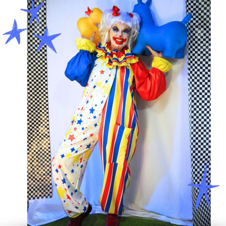 Op de afbeelding: Een clownskostuum bestaande uit een witte jumpsuit met een kleurrijk sterrenpatroon aan de ene kant en verticale rode, gele en blauwe strepen aan de andere kant. De outfit bevat een ruchekraag, lange mouwen en een bijpassende witte pruik.