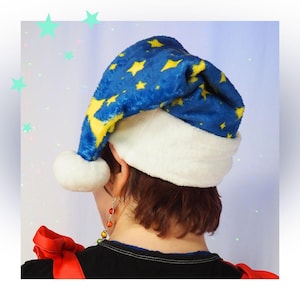 Puede incluir: Un gorro de felpa azul con un patrón de estrellas amarillas, un borde de peluche blanco y un pompón blanco en la punta. El gorro se lleva puesto en la cabeza, con la tela suave ligeramente inclinada hacia un lado, destacando el contraste de colores.