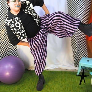 Peut inclure: Une chemise noire à manches courtes avec un motif graphique blanc composé d'yeux de différentes tailles, associée à un pantalon ample à rayures verticales alternées noires et violet clair. Le pantalon possède une taille élastique.