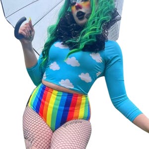 May include: A blue crop top with white cloud designs and long sleeves, paired with high-waisted rainbow shorts. The shorts feature vertical stripes in various colours. The outfit is completed with fishnet stockings.