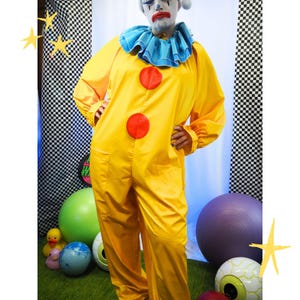 Puede incluir: Un mono de payaso de color amarillo brillante con mangas largas, dos grandes botones circulares rojos en la parte delantera y un cuello azul con volantes. La prenda tiene un corte holgado con bolsillos laterales visibles y se lleva con maquillaje blanco.