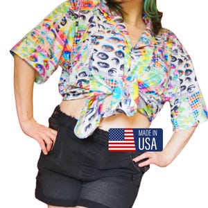 May include: A colorful button-down shirt with an eye pattern is tied at the waist. The shirt features a vibrant mix of colors and patterns. Black denim shorts are also visible. A "Made in USA" sticker is on the bottom right.