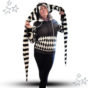 May include: Black and white zip-up hoodie with a unique design. The hood and long extensions feature bold black and white stripes. The body of the hoodie has a diamond pattern and circular designs. The person is wearing black leggings.