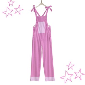 May include: A light pink overall jumpsuit with a square patch on the back and white trim at the bottom of the legs. The straps are tied at the shoulders. The jumpsuit is hanging on a white hanger.