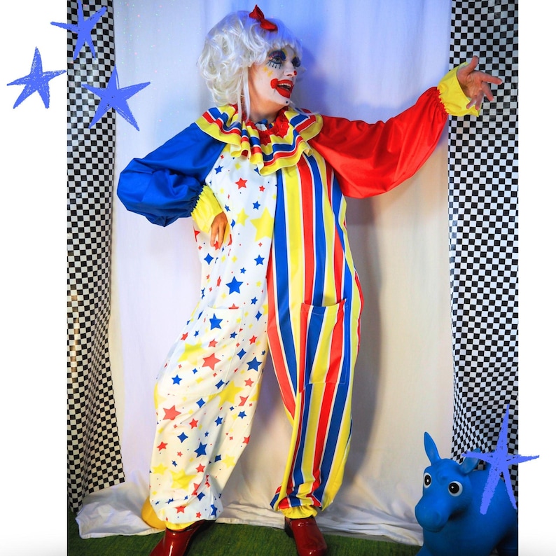 Op de afbeelding: Een clownskostuum bestaande uit een witte jumpsuit met een patroon van rode en gele sterren aan de ene kant en verticale rode, gele en blauwe strepen aan de andere kant. Het bevat een ruchekraag, lange mouwen met gele boorden en een witte pruik met een rode strik.