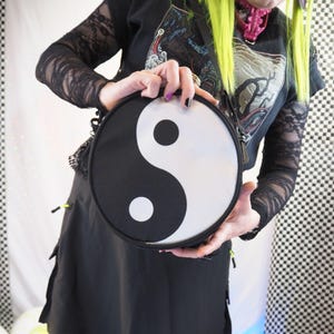 以下が含まれることがあります： 黒と白の陰陽のクロスボディバッグ。バッグは黒い外観で、白い陰陽のシンボルが付いています。バッグは黒い服と白黒のチェック柄の背景の前に保持されています。