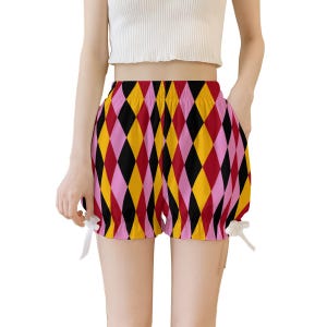 Könnte beinhalten: Hoch taillierte Shorts mit einem lebendigen Harlekin-Rautenmuster in abwechselnden Farben wie Rot, Gelb, Schwarz und Pink. Die Shorts verfügen über einen elastischen Bund und gerüschte Beinabschlüsse, die an beiden Seiten mit kleinen weißen Schleifen verziert sind.