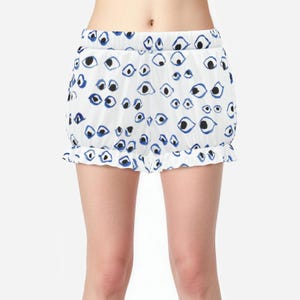May include: White shorts featuring a repeating pattern of hand-drawn blue and black eyes. The shorts have an elasticized waistband and a ruffled hemline along the bottom edge, creating a gathered, voluminous silhouette with a consistent print.