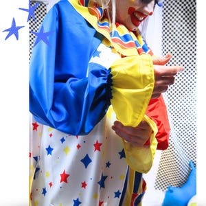 Op de afbeelding: Een clownskostuum met een witte basis en een patroon van blauwe en rode sterren. Het kledingstuk heeft lange mouwen met gele boorden, een ruchekraag met blauwe, gele en rode strepen, en effen blauwe en rode delen op de mouwen.