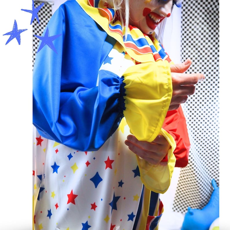 Op de afbeelding: Een clownskostuum met een witte basis en een patroon van blauwe en rode sterren. Het kledingstuk heeft lange mouwen met gele boorden, een ruchekraag met blauwe, gele en rode strepen, en effen blauwe en rode delen op de mouwen.