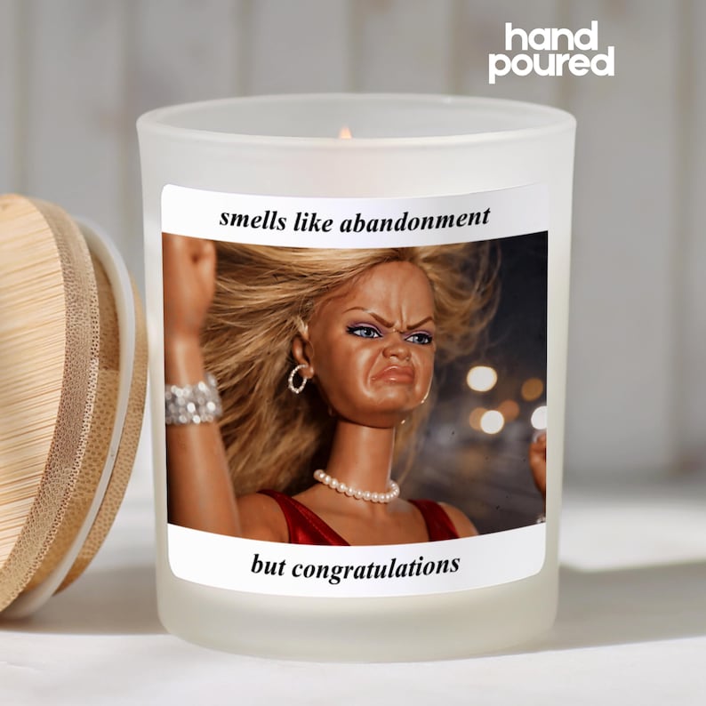 K&ouml;nnte beinhalten: Eine Kerze aus mattiertem Glas mit einem Holzdeckel. Die Kerze hat ein Etikett mit dem Text "smells like abandonment but congratulations" und ein Foto einer Puppe mit missmutigem Gesichtsausdruck. Die Worte "hand poured" stehen oben.