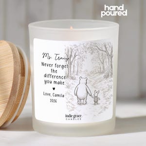 May include: A frosted glass candle with a wooden lid. The candle features a label with a black and white illustration of a bear and pig walking on a path. Text on the label reads "Ms. Tracy, Never forget the difference you make."