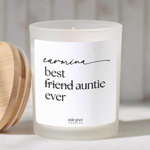 May include: A frosted glass candle in a cylindrical container with a bamboo lid. The white label features black text that reads: carmina, best friend auntie ever, and indie grace CANDLES. The candle has a visible flame at the top center.