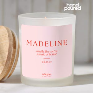 Pu&ograve; includere: Una candela in vetro smerigliato con coperchio in legno. La candela ha un'etichetta rosa con il nome "MADELINE" in rosso, la frase "smells like you're a maid of honor" e la data 04.05.27. Le parole "hand poured" sono in alto.