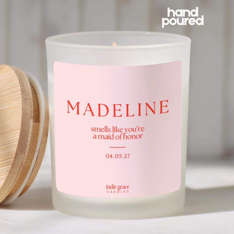 Pu&ograve; includere: Una candela in vetro smerigliato con coperchio in legno. La candela ha un'etichetta rosa con il nome "MADELINE" in rosso, la frase "smells like you're a maid of honor" e la data 04.05.27. Le parole "hand poured" sono in alto.