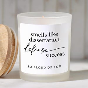 May include: A frosted glass candle in a cylindrical container with a bamboo lid. The white label features black text that reads: smells like dissertation defense success SO PROUD OF YOU. The candle has a single wick centered in the wax.