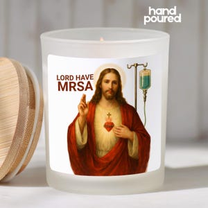 May include: A frosted glass candle with a wooden lid. The candle features a label with an image of Jesus and the text "LORD HAVE MRSA". The candle is hand poured.
