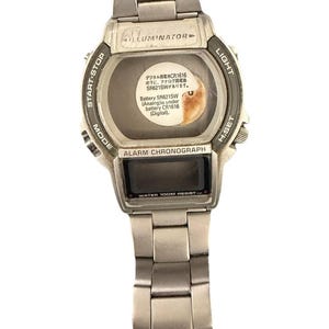 May include: A silver-tone metal wristwatch with a link bracelet. The watch face features the text ILLUMINATOR, ALARM CHRONOGRAPH, and WATER 100M RESIST. A central sticker provides battery information in Japanese and English text.